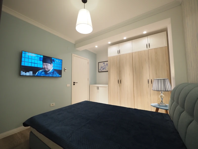 Compact apartment bedroom interior with bed, wardrobe, TV, and light wood finishes